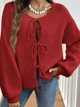 Woman wearing a red tie-front long sleeve knit cardigan with three bow ties paired with blue jeans.