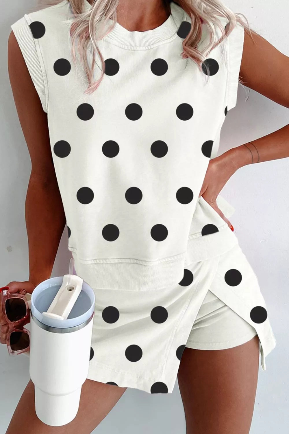 Person wearing a white sleeveless top with black polka dots and matching shorts, holding a white tumbler.