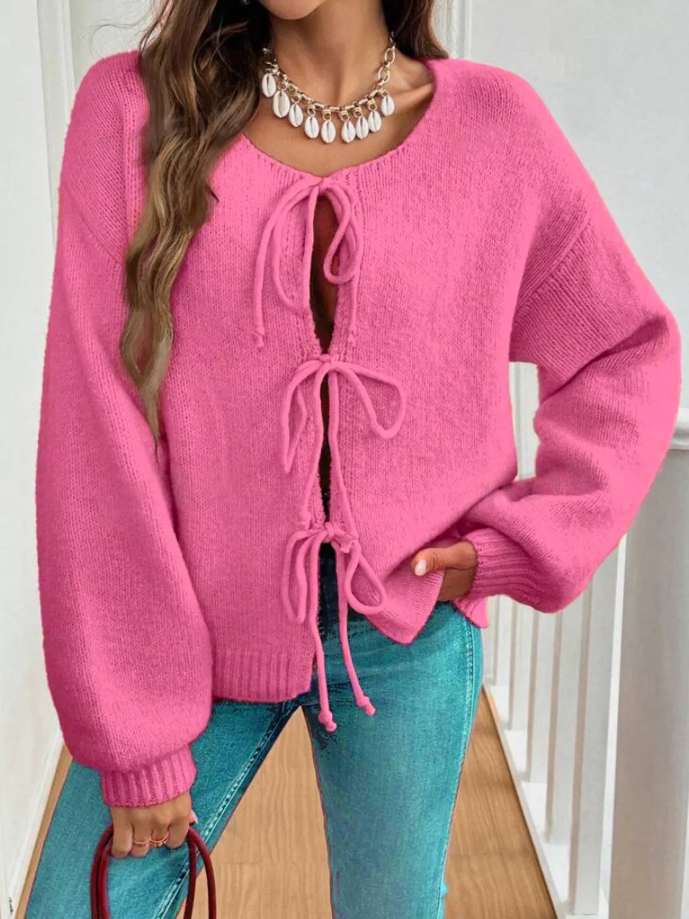 Woman wearing a rose-pink tie-front long sleeve knit cardigan with three bow ties paired with blue jeans.