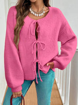 Woman wearing a rose-pink tie-front long sleeve knit cardigan with three bow ties paired with blue jeans.