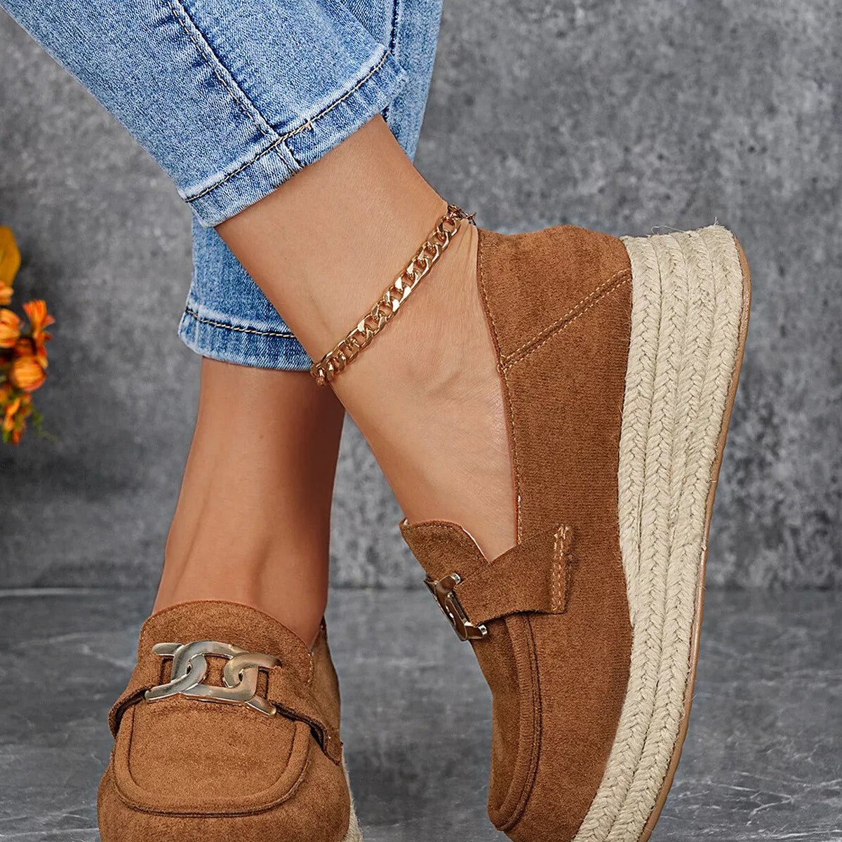 Brown suede loafers with chain detail worn with rolled-up jeans on a gray background