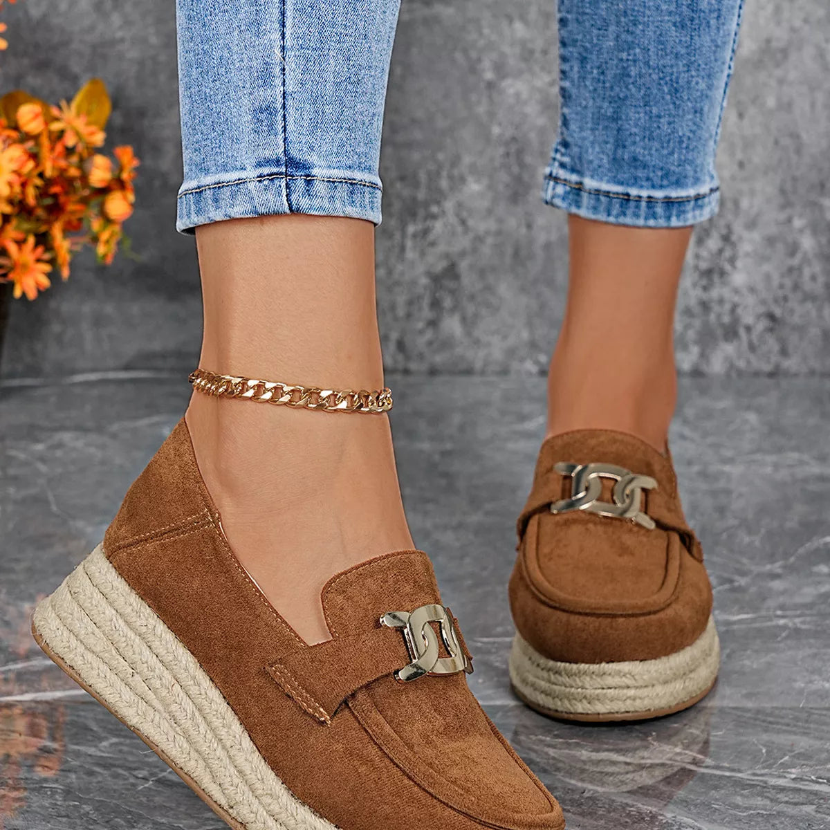 Brown loafers with gold buckles worn with blue jeans on a gray surface.