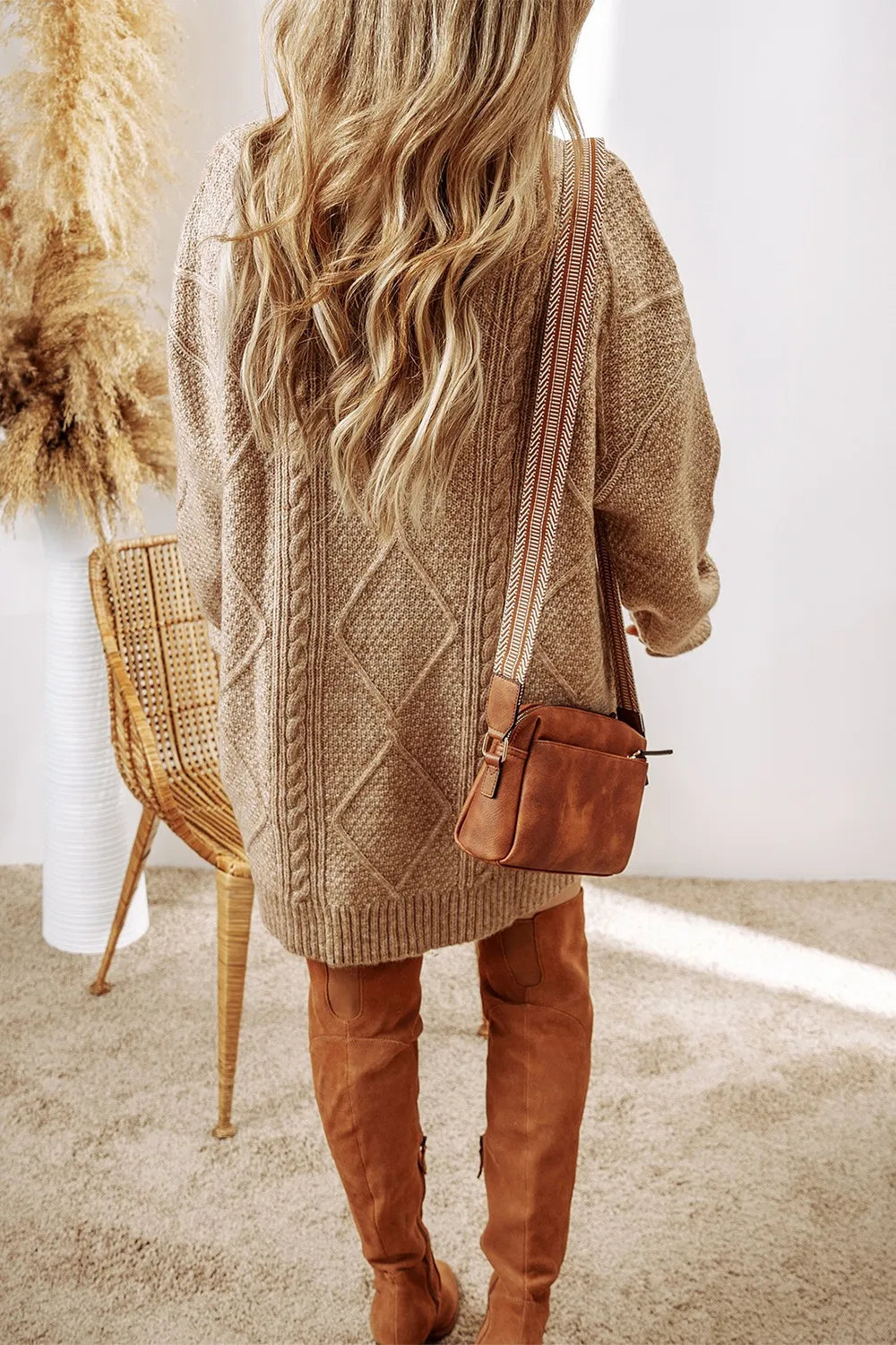 Woman wearing a tan cable knit long sleeve sweater dress styled with knee-high brown boots and layered gold necklaces.