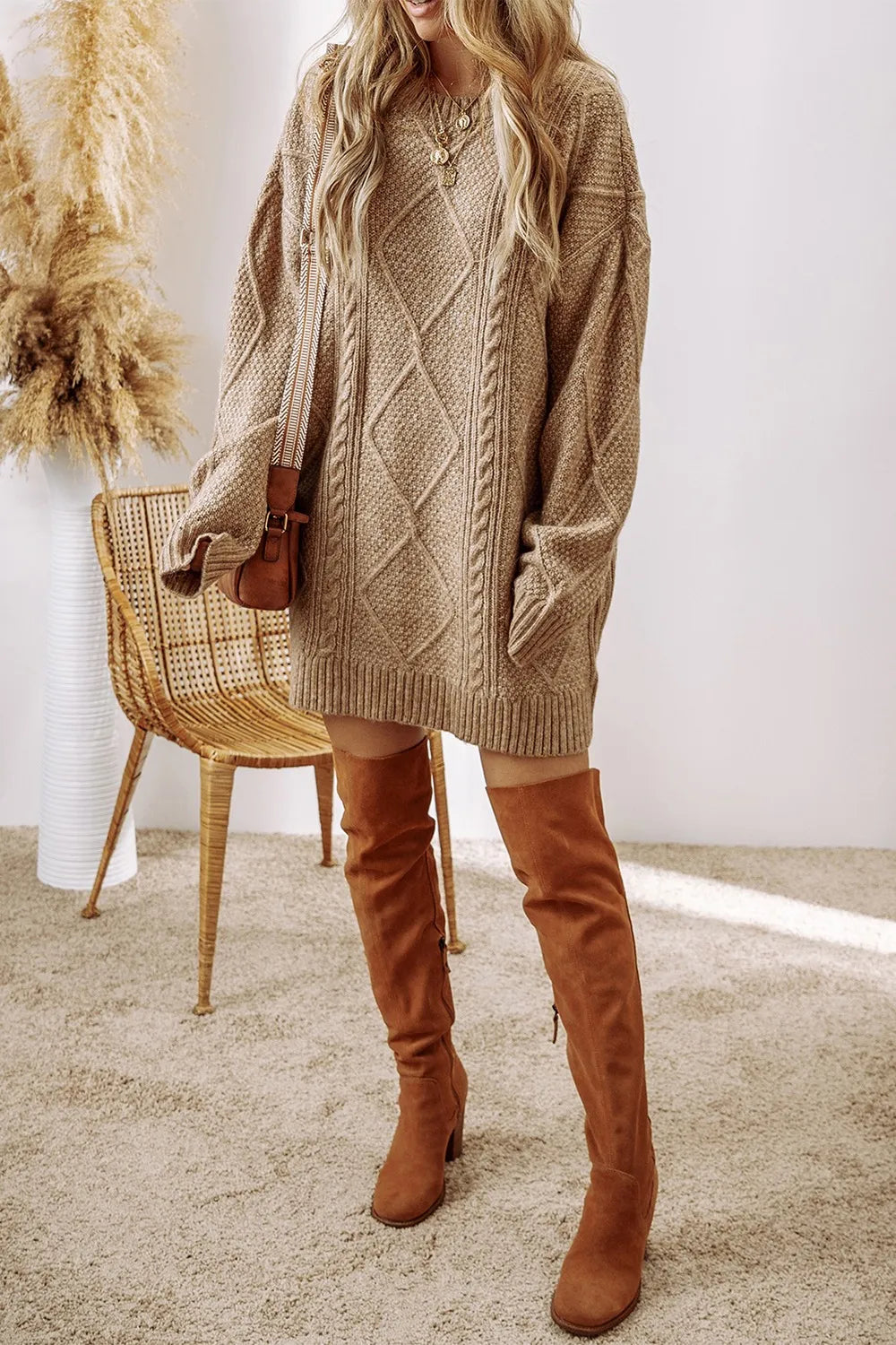 Woman wearing a tan cable knit long sleeve sweater dress styled with knee-high brown boots and layered gold necklaces.