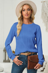Woman wearing a cerulean textured round neck long sleeve blouse with dark-wash jeans, brown clutch and cream hat.