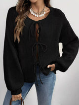 Woman wearing a black tie-front long sleeve knit cardigan with three bow ties paired with blue jeans.