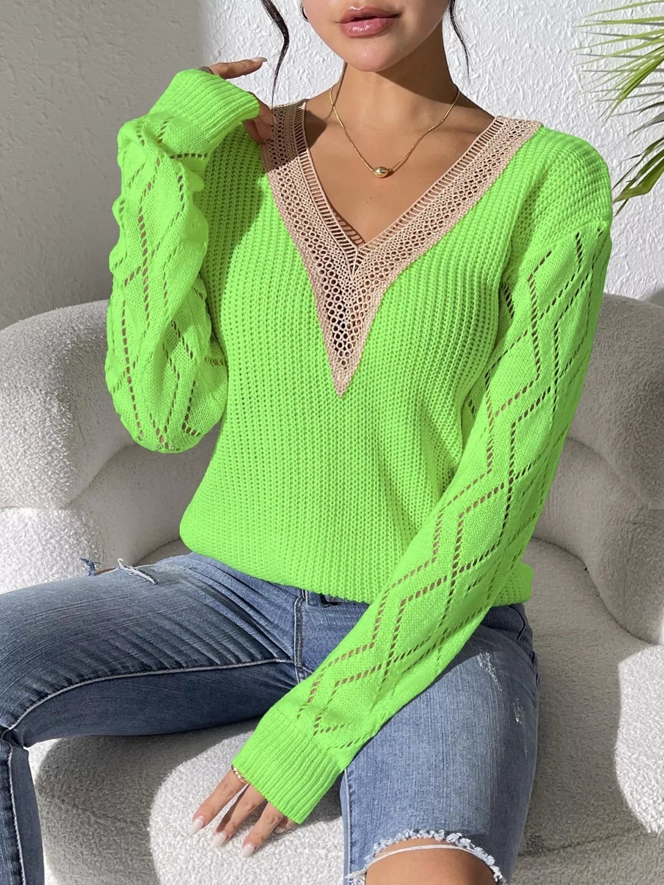 Woman wearing a green V-neck hollow knit sweater with contrasting neckline, openwork long sleeves, and blue jeans.
