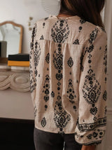 Woman wearing a beige and black vintage print long-sleeve blouse with dark denim jeans, a brown belt, and statement jewelry.