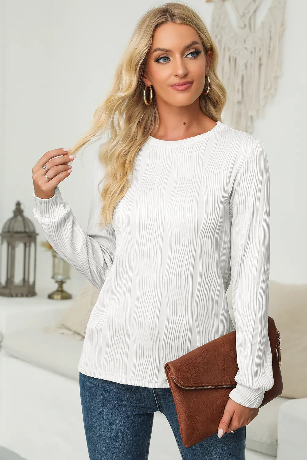 Woman wearing a white textured round neck long sleeve blouse with dark-wash jeans, tan belt and brown clutch.