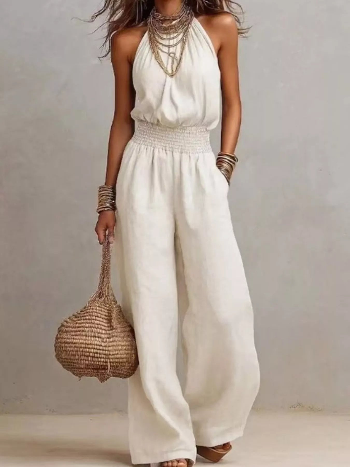 Woman wearing a white halter-neck wide-leg jumpsuit with a smocked waist, layered gold jewelry, and bracelets, styled for an elegant evening look.