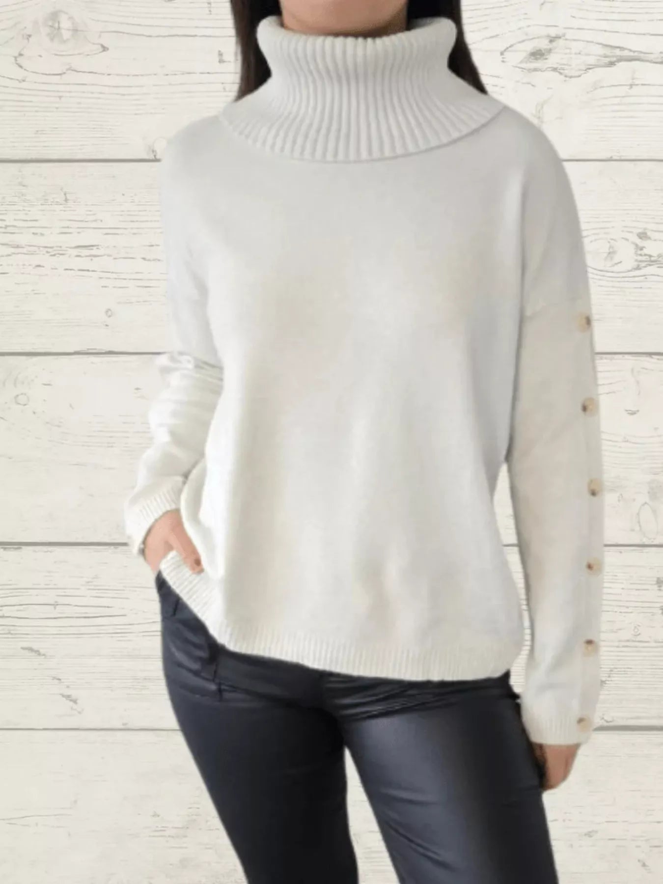 Woman wearing a white ribbed turtleneck sweater with decorative button sleeves and jeans.