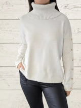 Woman wearing a white ribbed turtleneck sweater with decorative button sleeves and jeans.