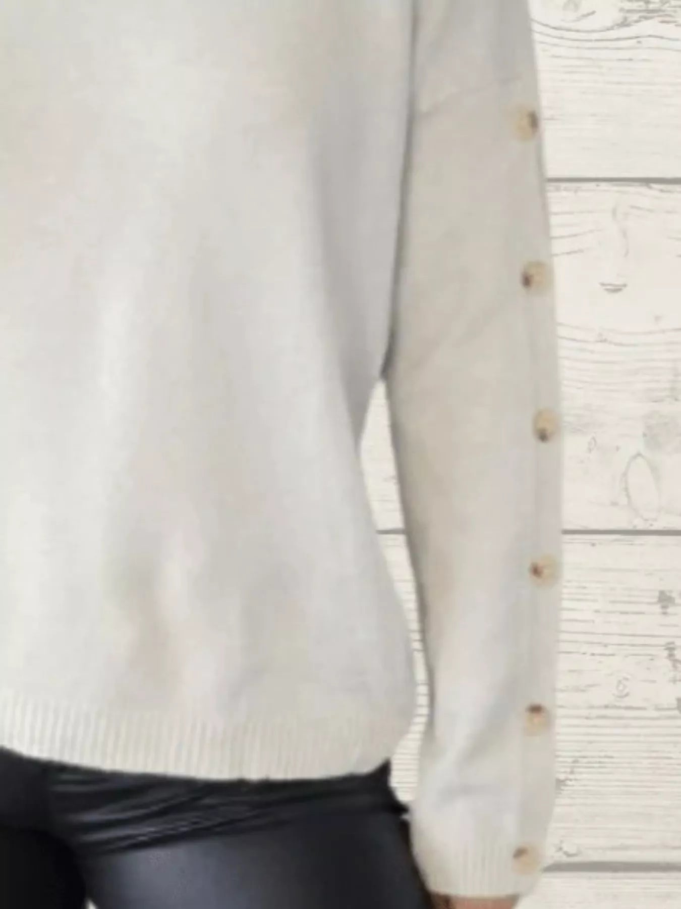 Woman wearing a white ribbed turtleneck sweater with decorative button sleeves and jeans.