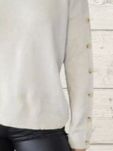 Woman wearing a white ribbed turtleneck sweater with decorative button sleeves and jeans.