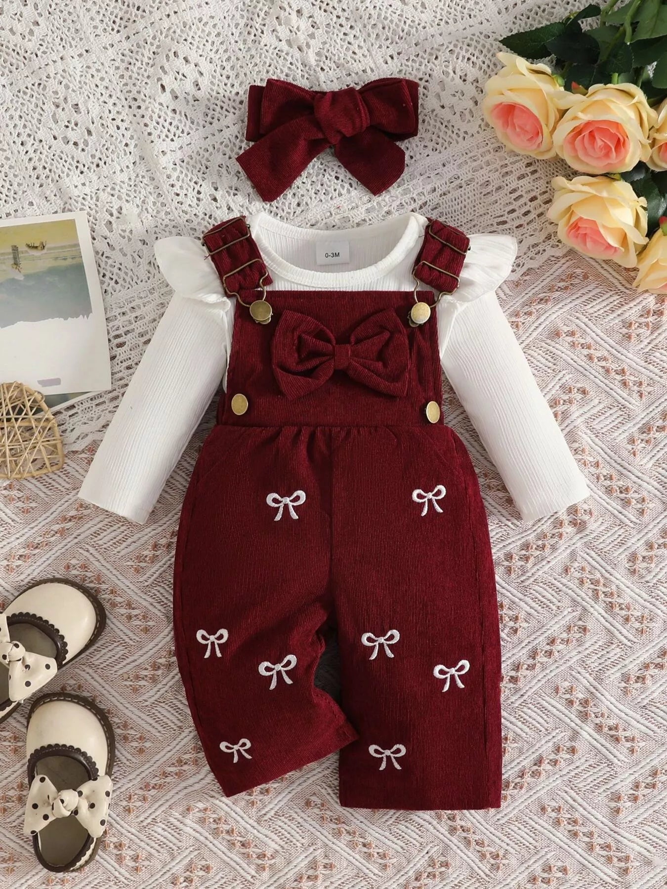 Baby girl three-piece outfit featuring a cream ribbed long-sleeve bodysuit with ruffled shoulders, wine red bow-embroidered overalls and matching wine-red bow headband.