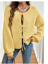 Woman wearing a yellow tie-front long sleeve knit cardigan with three bow ties paired with blue jeans.